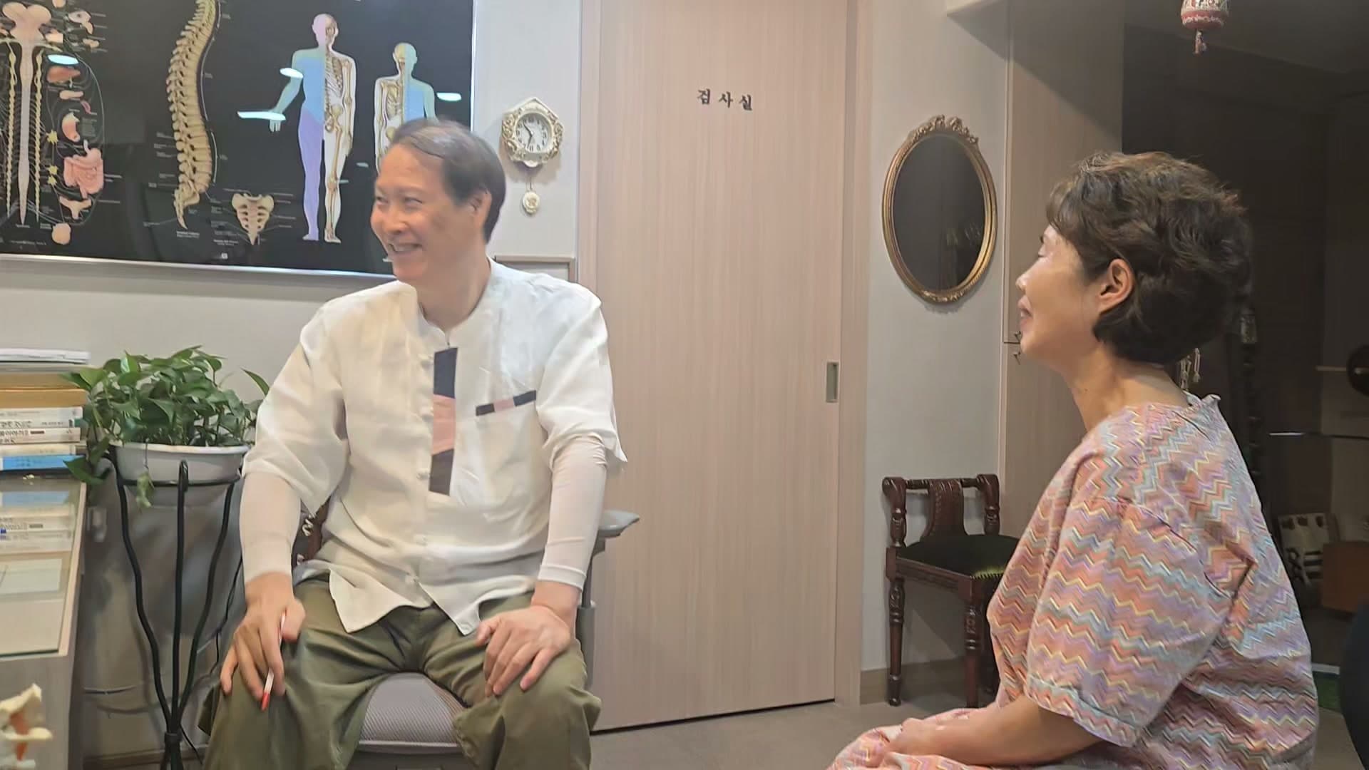 진료실에서 의료진과 환자가 허리 통증과 걸을 때 앞으로 숙여지는 자세에 관해 이야기하는 장면